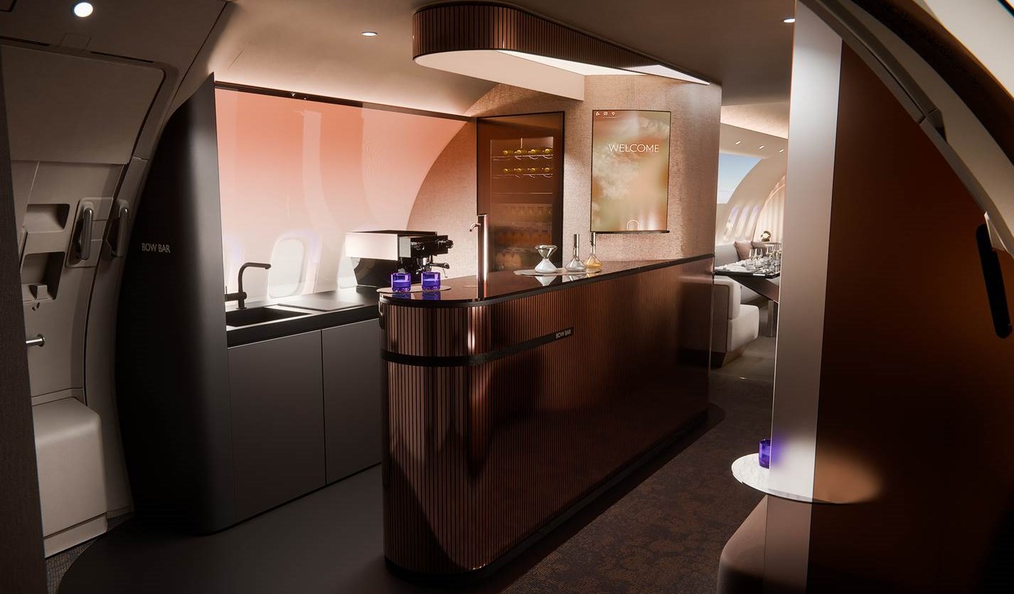 The BOW. Der Barbereich an Bord zeigt eine Holztheke mit Espressomaschine und Weinkühlschrank. Digitaler Bildschirm mit Willkommensgruss. (Credits: Designworks und Lufthansa Technik)